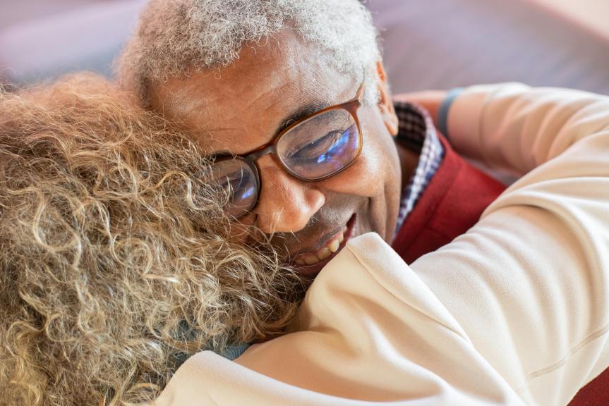 Close-up of an older man wearing glasses smiling as he hugs another person with curly hair, showing warmth and affection.