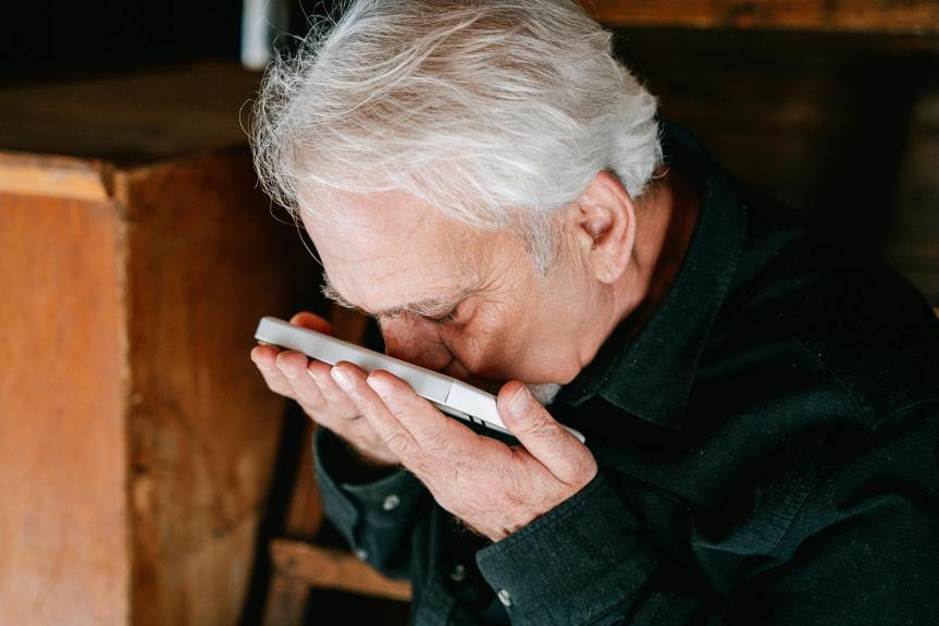 Older adult with white hair sits indoors kissing a picture, holding it close to his face in a quiet, reflective moment