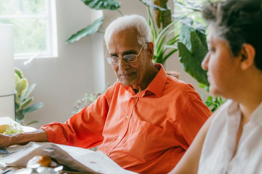 Older man in an orange shirt sits at a table reading a newspaper while another person sits nearby in a bright, plant-filled home.