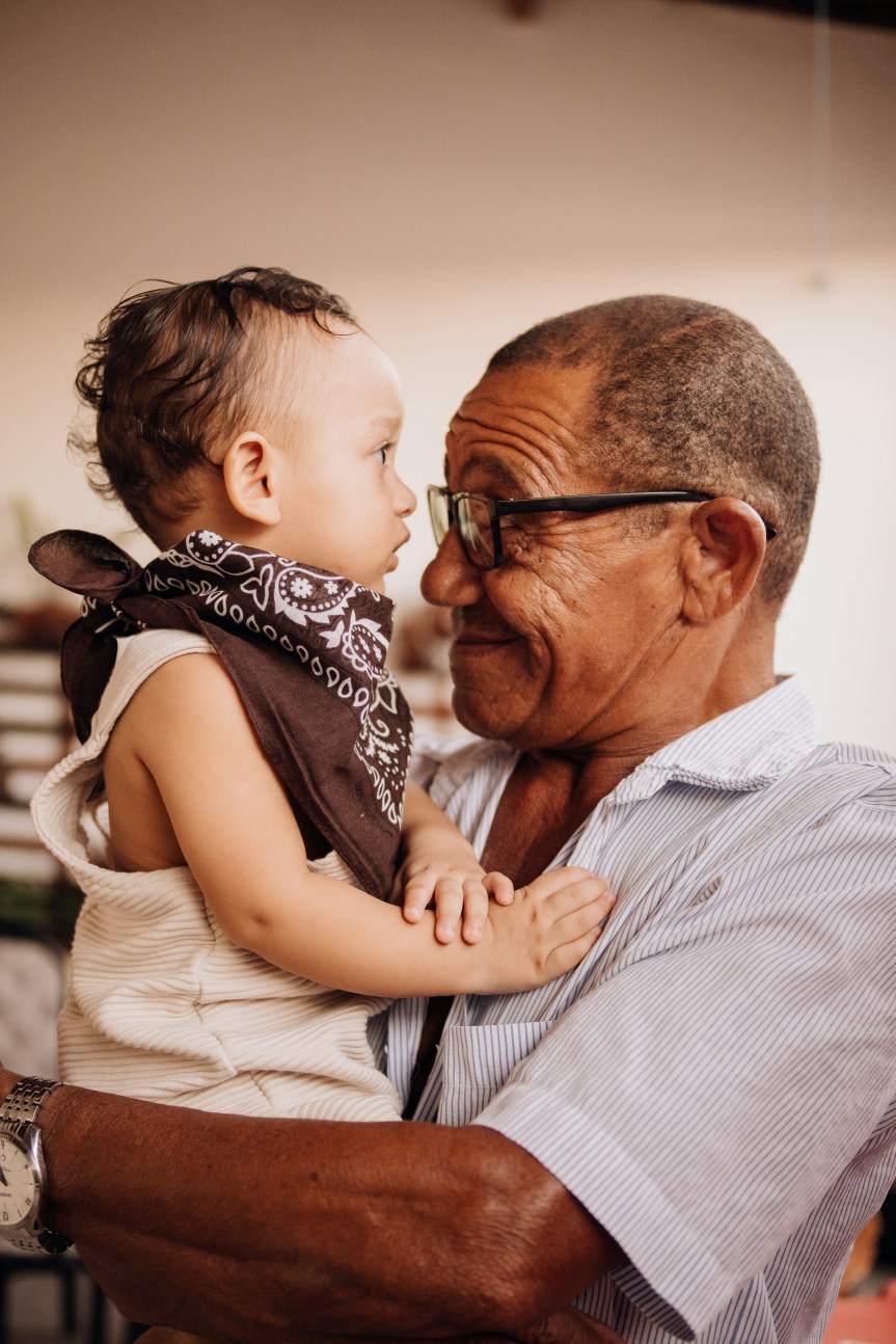 Photo of an elderly black man holding a baby wearing a brown bandana around his neck. The elderly man wears black glasses and a striped blue and white collared shirt.