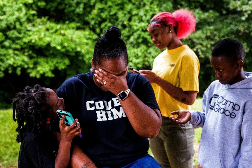 A woman sits outdoors covering her face with one hand while three children stand close around her, capturing a moment of stress and overwhelm in a busy family routine.