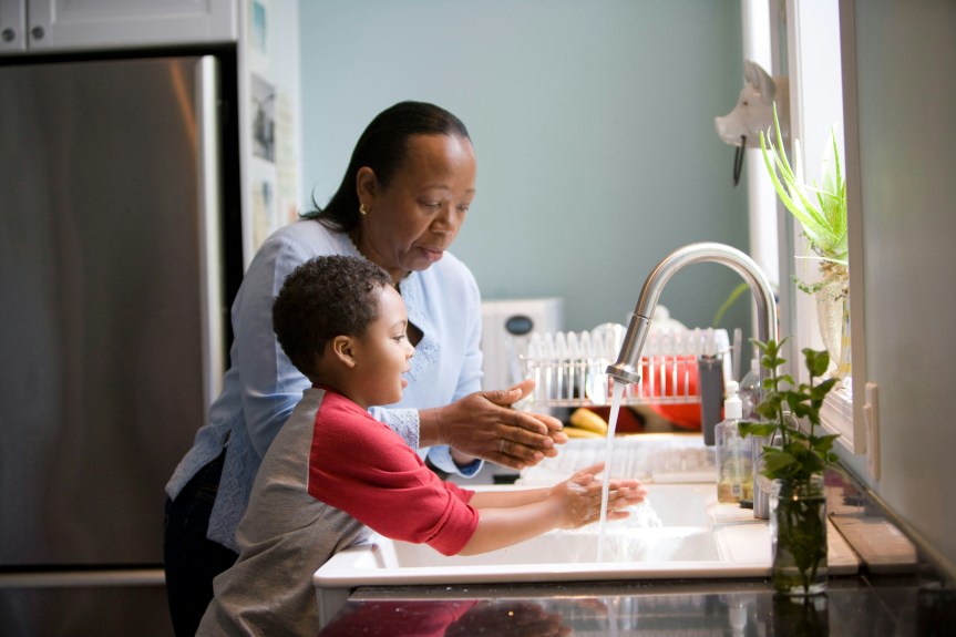 A woman stands beside a young child at a kitchen sink, guiding them as they wash their hands together.