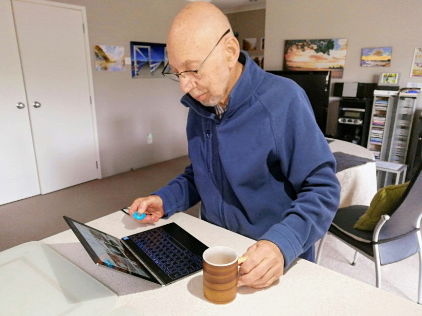 Photo of an elderly man standing at a desk in an office space on a laptop, with a cup of coffee in his left hand.